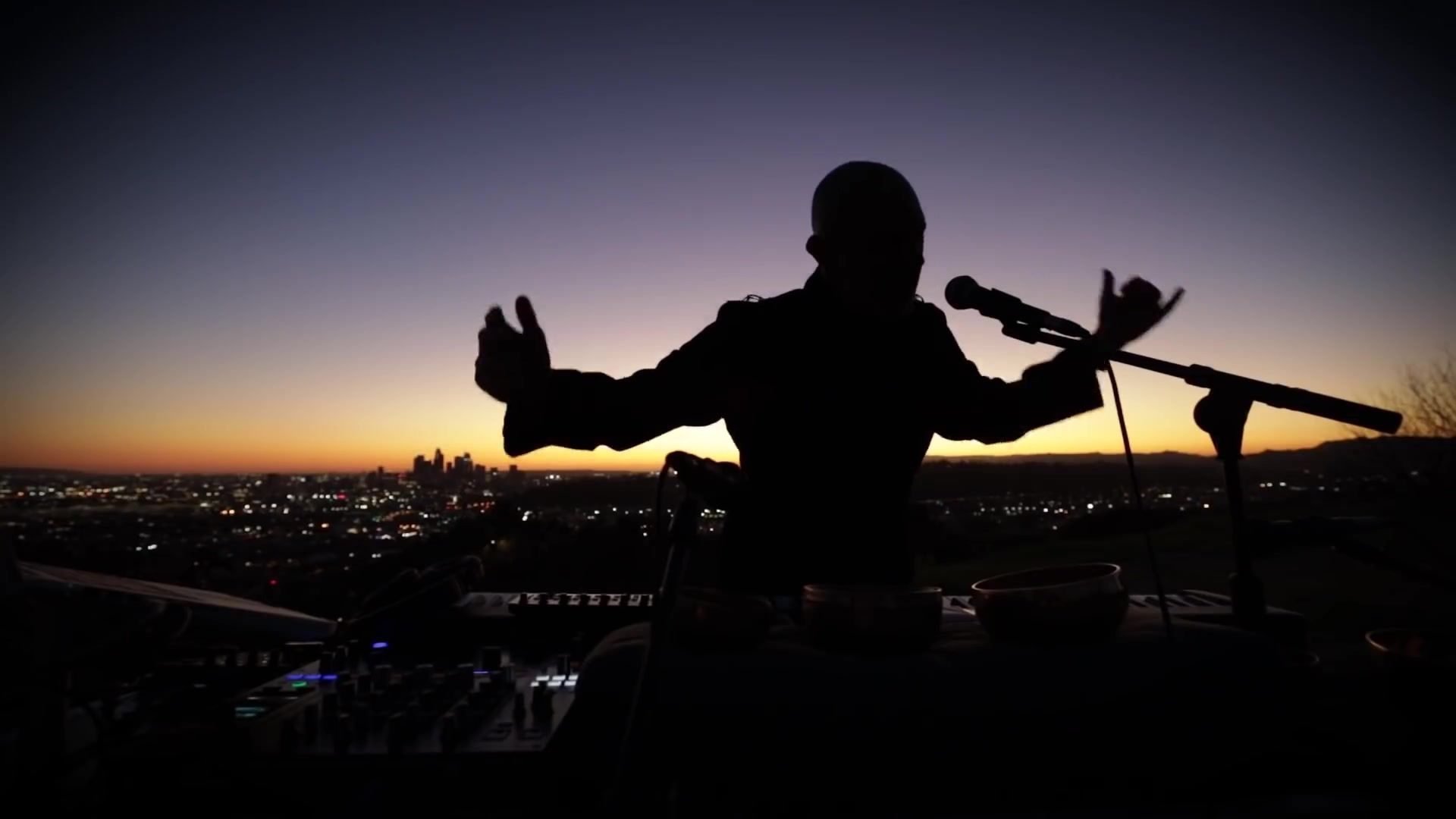 Reza Safinia in silhouette with arms outstretched, performing on a hilltop at dusk above Los Angeles
