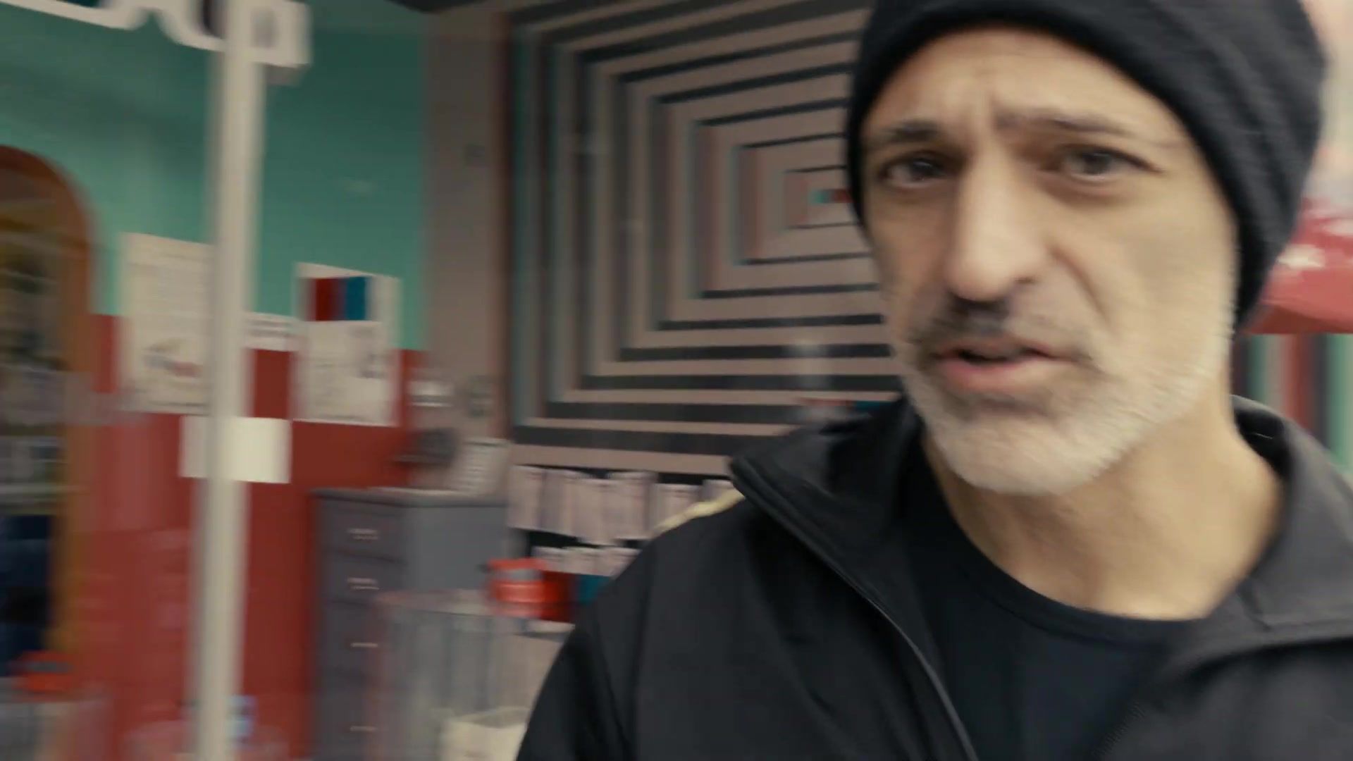 Reza Safinia in a black beanie and track jacket, singing to camera in a Los Angeles shop